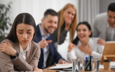 Female employee looking concerned at her desk, while colleagues talk behind her back.