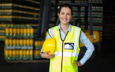 Female supervisor in hard hat holding clipboard while trucks load in the background, symbolizing logistics and safety.