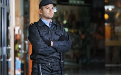 A security guard standing outside an office building during evening hours, suggesting long shifts and wage concerns.