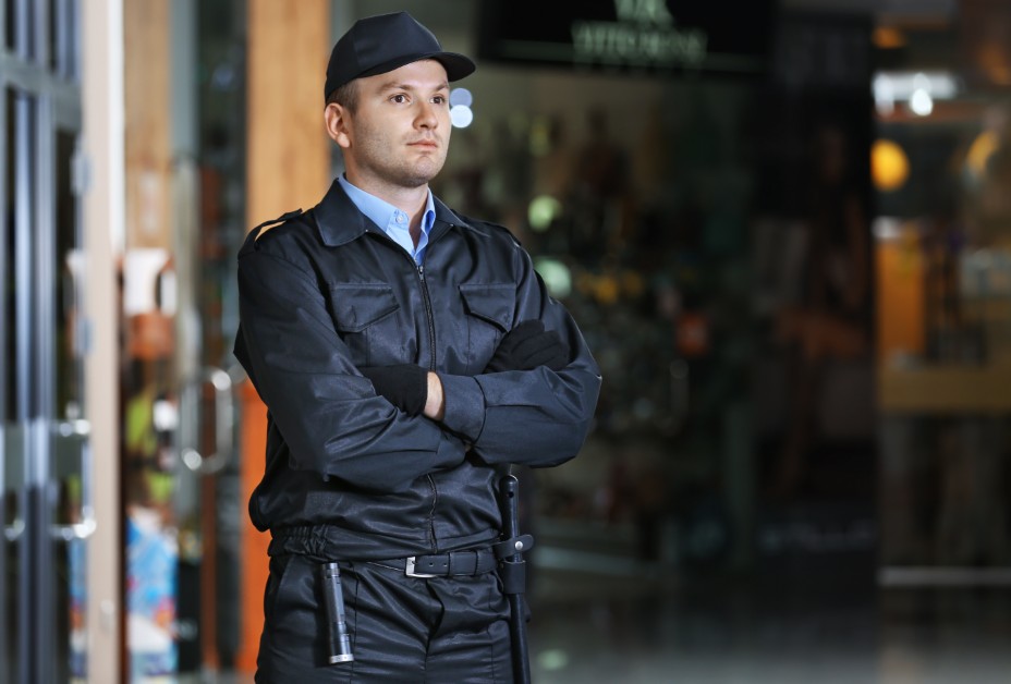 A security guard standing outside an office building during evening hours, suggesting long shifts and wage concerns.