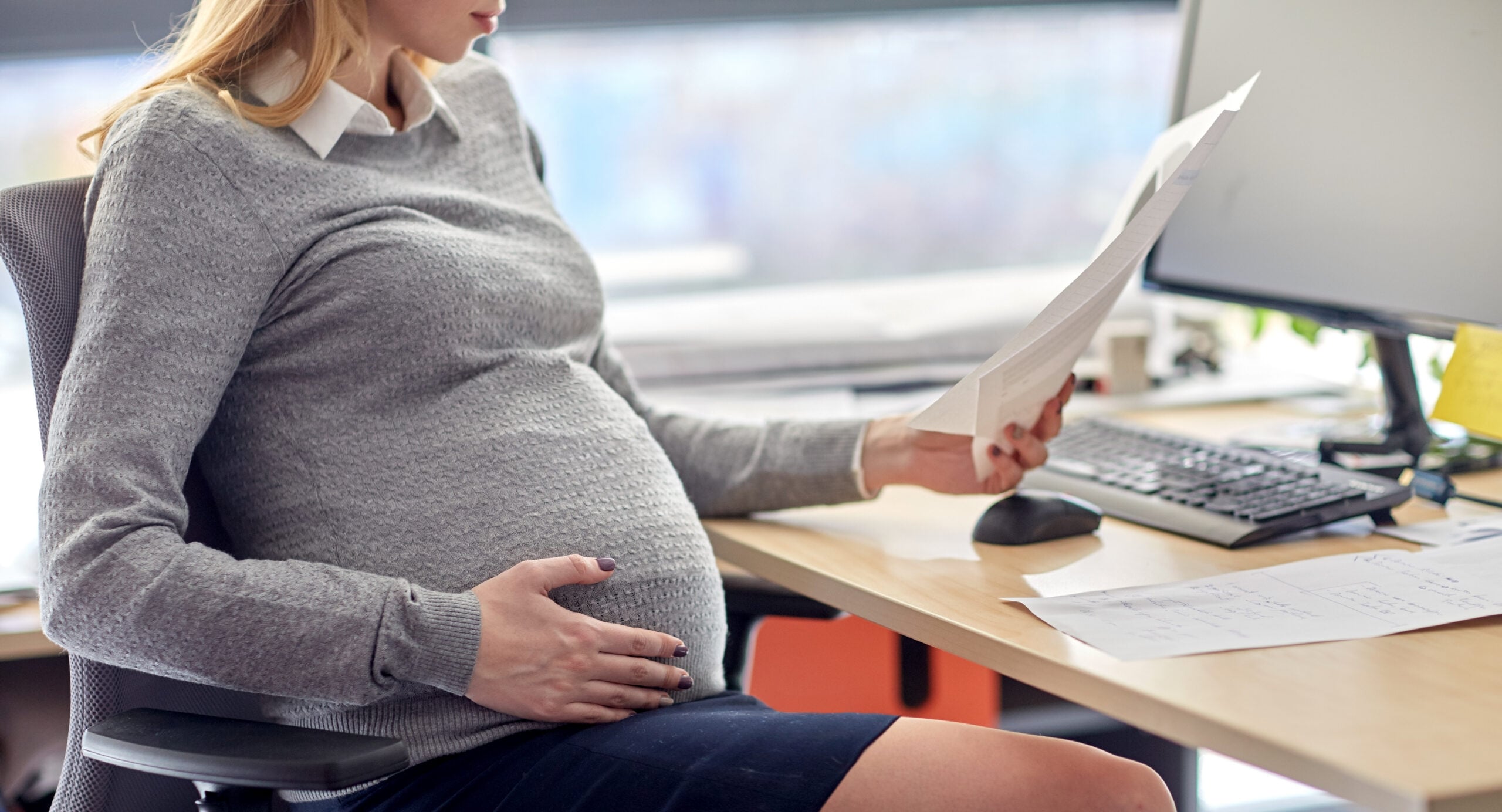 Pregnant employee reviewing workplace emails and notes at desk 